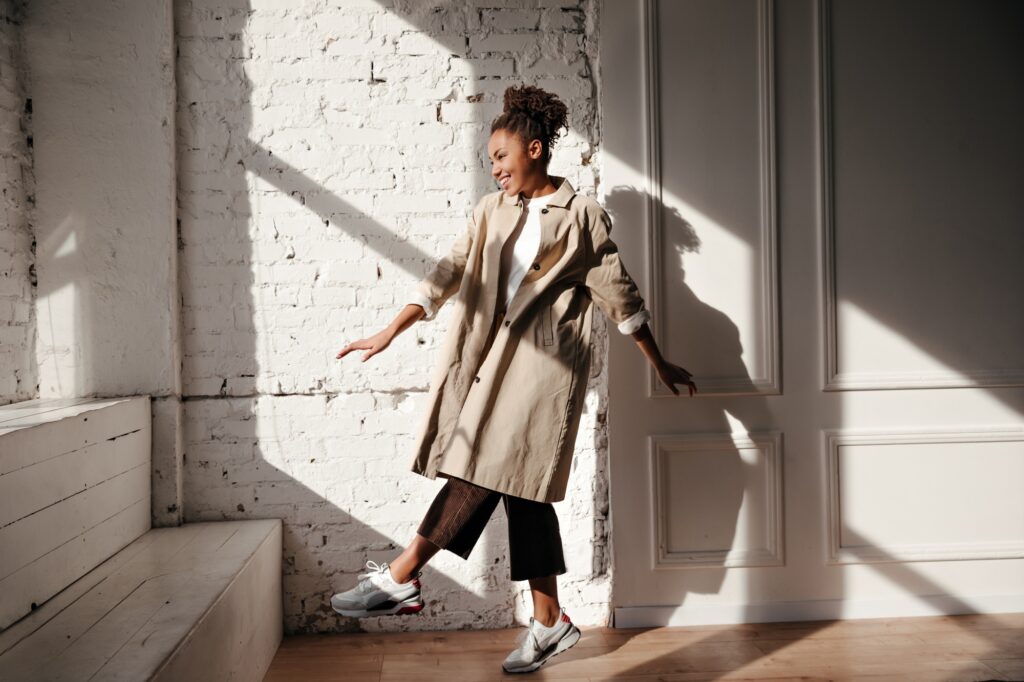 Full length view of pleasant black woman in trench coat. Laughing african american girl dancing und